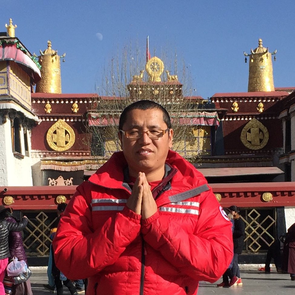 portrait of man with glasses posing with prayer hands in front of monastery