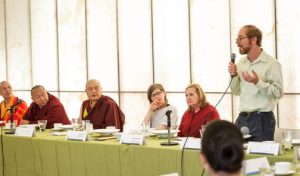 man speaking into microphone at a table alongside women and monks