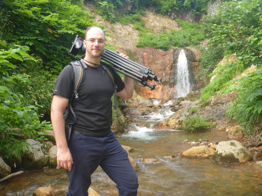 man posing with tripod over shoulder in front of waterfall