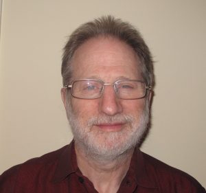 headshot of gray haired bearded man with glasses smirking